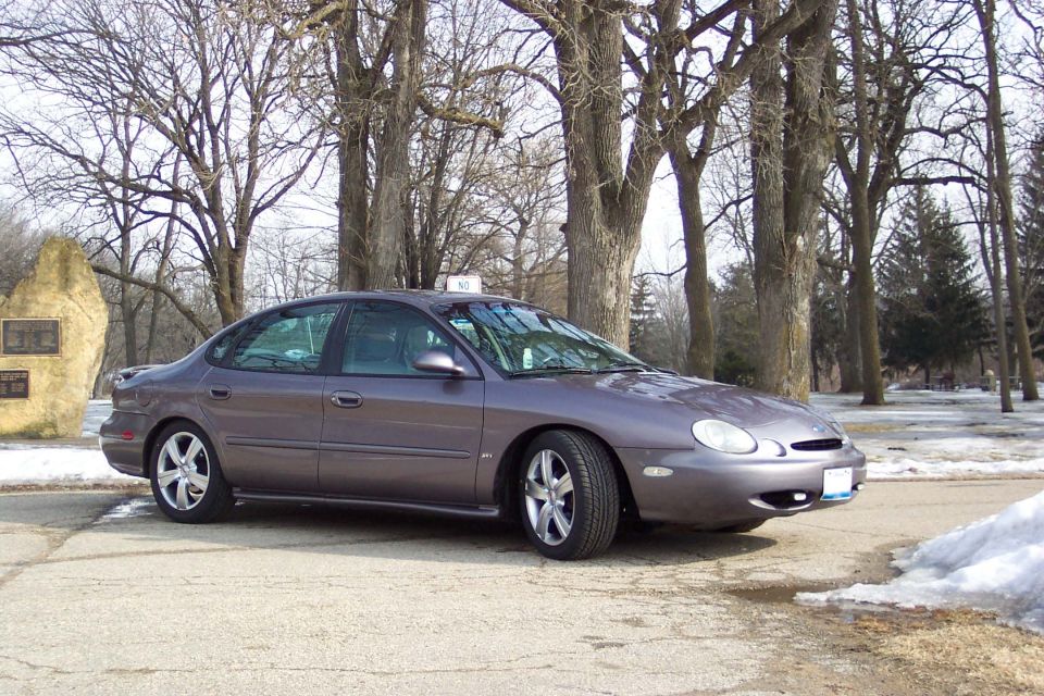 Gray Ford Taurus parked on a paved area with trees and snow in the background