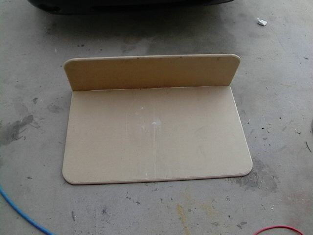 Cardboard template with a raised back edge on a concrete floor, with a blue hose and part of a black car bumper visible