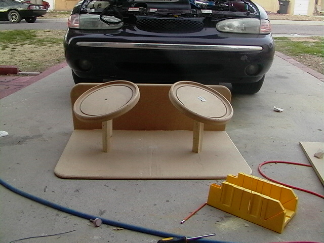 MDF speaker box with oval rings being built in front of a dark Ford Taurus with hood open