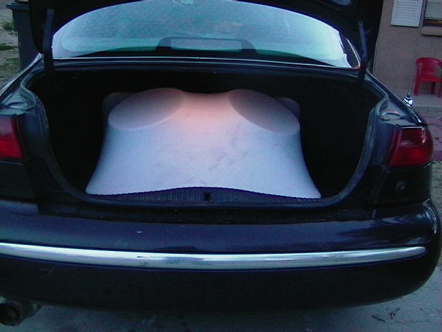 Black sedan's open trunk reveals a large, white, curved object. Chrome trim on bumper