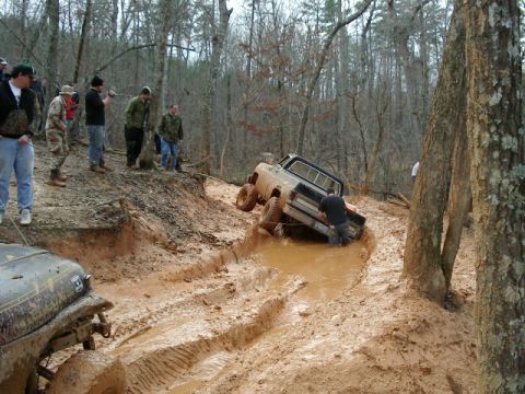 1985 Ford Bronco - Off-roaders navigate a challenging muddy terrain in a wooded area.
