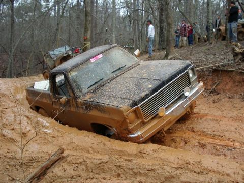 1985 Ford Bronco - This pickup truck is stuck deep in the mud, with onlookers gathered in the background.