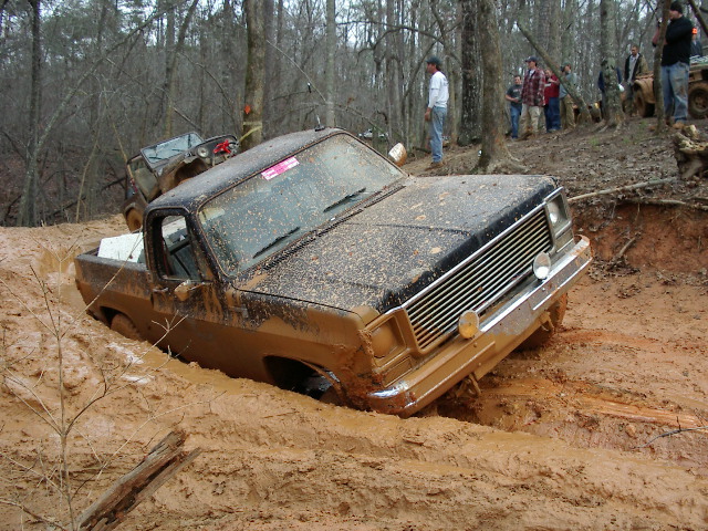 Muddy pickup truck stuck in a ditch with people watching. Another vehicle is behind it