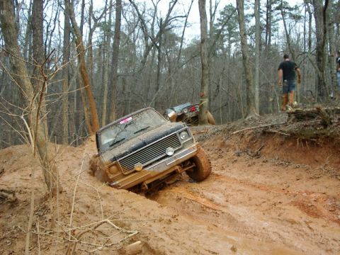 1985 Ford Bronco - A mud-caked square-body full-size 4x4 SUV with a chrome slat grille, dual round headlights and lifted suspension on mud-terrain tires climb…