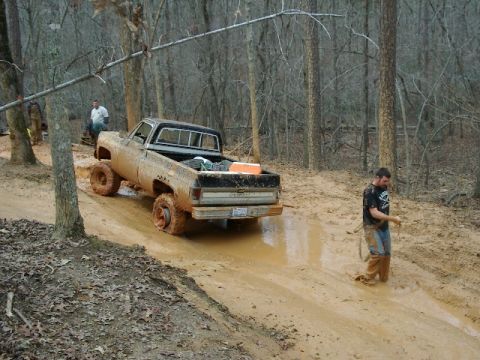1985 Ford Bronco - Rear three-quarter view of a mud-caked lifted Chevrolet square-body pickup with a black cab, orange cooler and gear in the bed, backing thr…