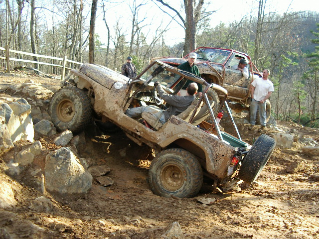 Mud-covered Jeep Wrangler with open top navigating rocky terrain, driver visible