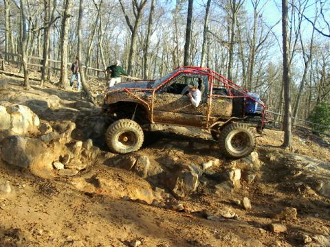 1985 Ford Bronco - Side view of a mud-covered, chopped-roof mini truck buggy with a red exposed tube roll cage crawling down a rocky ledge on a wooded off-roa…