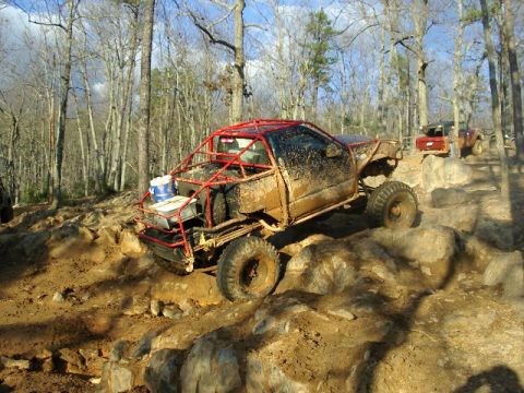 1985 Ford Bronco - Rear three-quarter view of a mud-splattered stripped mini truck with a red exposed tube cage, no doors or bed sides, and a blue bucket stra…