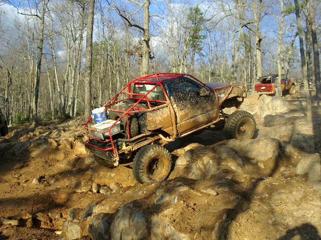 Muddy stripped mini truck with red tube cage crawling over rocks on a wooded trail