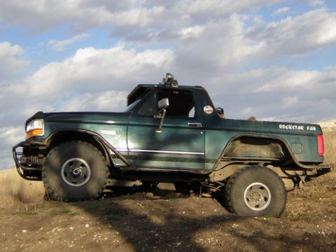 1995 Ford Bronco - Side profile of a dark green Ford Bronco with the roof removed, parked on dirt under a big cloudy sky. It has a front brush guard, rocker g…