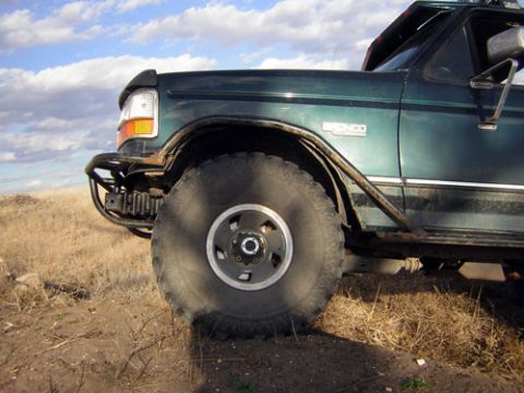 1995 Ford Bronco - Low front-quarter view of a lifted dark green Ford Bronco parked in dry grass under a blue sky with scattered clouds, cropped to show the f…