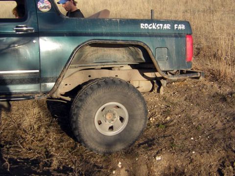 1995 Ford Bronco - Passenger-side rear quarter of a dark green Ford Bronco parked in dry grass, showing a cut rear fender with tube rock slider and exhaust ex…