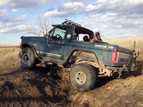 1995 Ford Bronco - Dark green lifted Ford Bronco with its top and doors removed, flexing over a dirt berm in a dry grass field, shot from the driver's-side re…