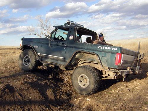 Green doorless lifted Ford Bronco flexing over a dirt berm in a field, rear three-quarter view
