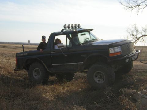 1995 Ford Bronco - A black Ford Bronco with off-road modifications is driven through a field by two individuals.