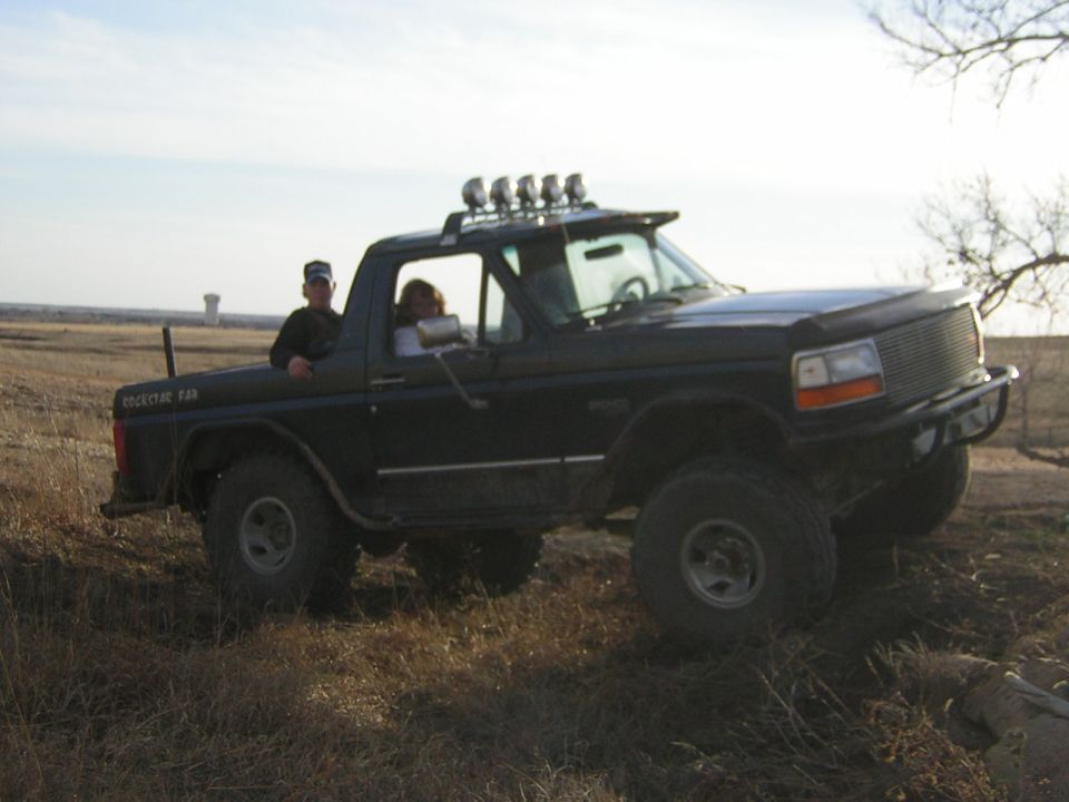 Black Ford Bronco with off-road tires and light bar, driven by two people in a dry field