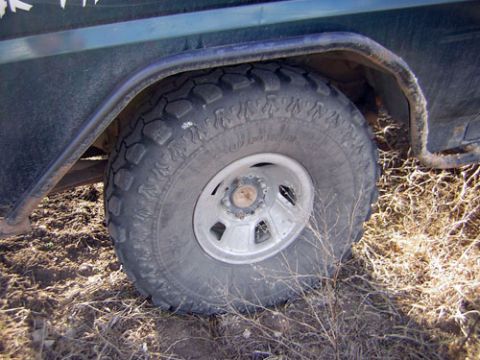 1995 Ford Bronco - Close-up of the front wheel of a dark green truck sitting in dry grass and dirt, fitted with a mud-terrain tire on a five-spoke steel-look…