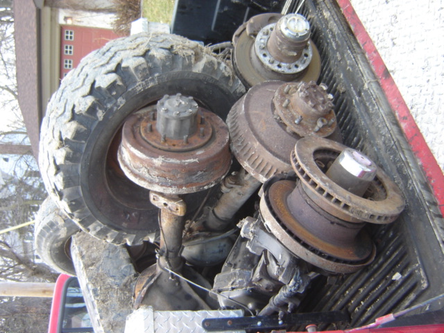 Pickup truck bed loaded with rusty axle parts and a large tire