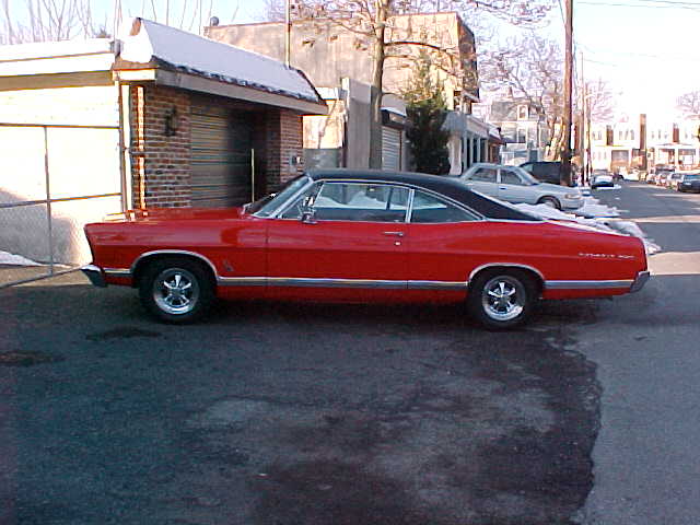 Red vintage coupe with black roof parked in profile by a brick garage.