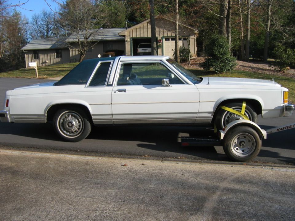 White Ford Crown Victoria coupe loaded on a tow dolly, side view in a residential setting.