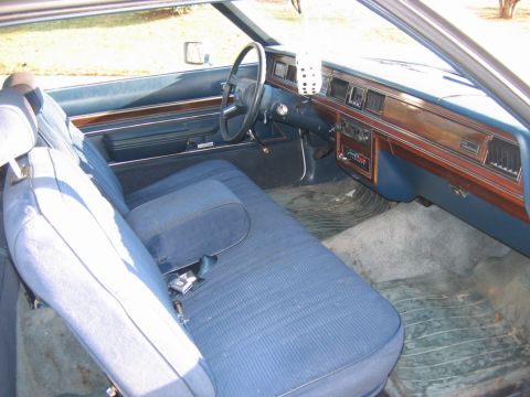 1985 Ford Crown Victoria LTD - The interior of a blue car features ribbed cloth seats, a wood-trimmed dashboard, and a dice air freshener hanging from the rearview mirror.