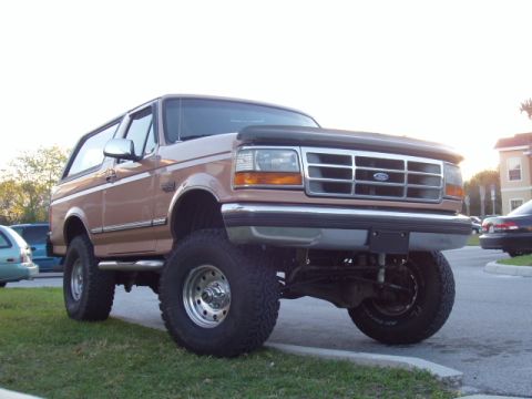 1994 Ford Bronco - This lifted tan Ford Bronco has a chrome grille and aftermarket wheels, parked on grass next to a street.