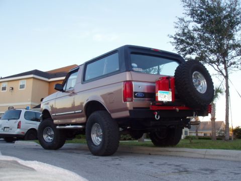 1994 Ford Bronco - This lifted Ford Bronco is parked on a street with a building and trees in the background.
