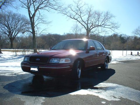 1999 Ford Crown Victoria - Maroon sedan parked on wet asphalt with snow in the background. The car appears to be a Ford Crown Victoria, likely from the late 1990s.