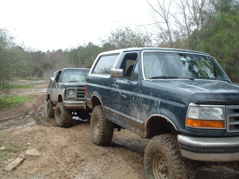 1993 Ford Bronco - Two lifted SUVs navigate a muddy trail, kicking up dirt as they go.