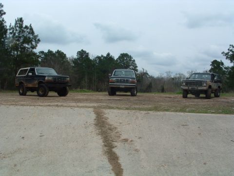 1993 Ford Bronco - Three SUVs are parked on a dirt lot under a cloudy sky, with the leftmost vehicle being a dark 1992 Ford Bronco.