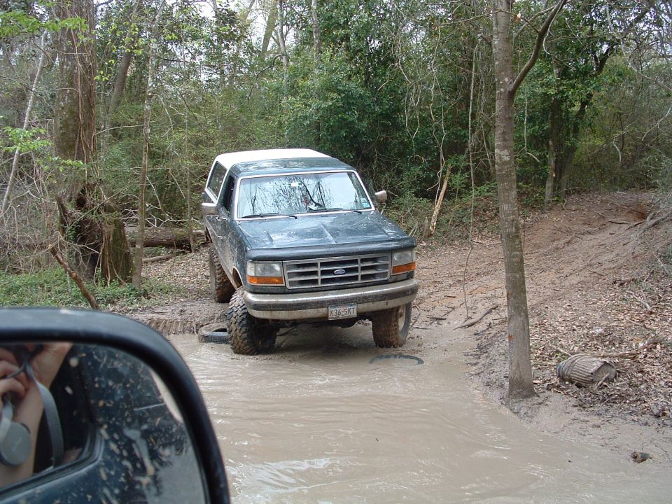 1993 Ford Bronco driving through a muddy trail with trees on either side
