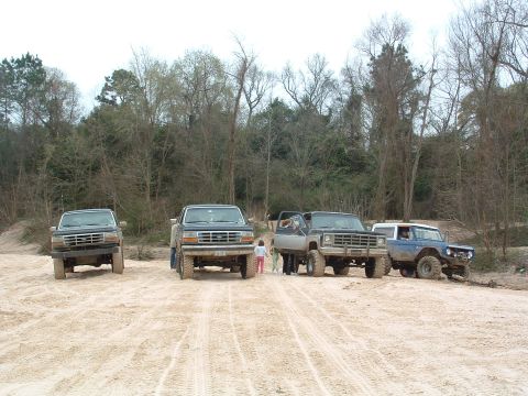 1993 Ford Bronco - Several off-road vehicles are parked on a dirt lot with trees in the background.