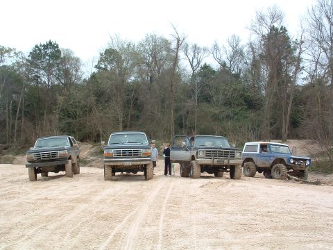 1993 Ford Bronco - Four off-road vehicles, including a 1993 Ford Bronco, are parked on a sandy terrain under an overcast sky.