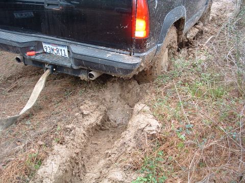 1993 Ford Bronco - A black SUV is deeply stuck in mud, with its rear tire submerged and a tow strap attached.