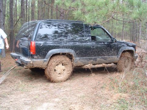 1993 Ford Bronco - A black SUV is deeply stuck in mud, with its tires caked in dirt. The vehicle appears to be a 1995 Chevrolet Tahoe.