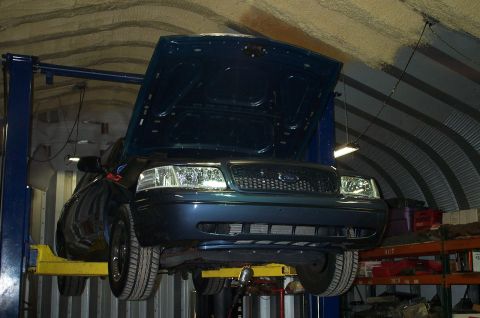 1998 Ford Crown Victoria - A dark blue/teal Ford sedan raised on a blue two-post lift with its hood open, viewed from the front in a curved corrugated-metal Quonset-s…