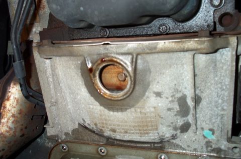 1998 Ford Crown Victoria - Close-up underside view of a metal transmission or oil pan with a round drain opening showing brown fluid staining and residue, mounted bel…