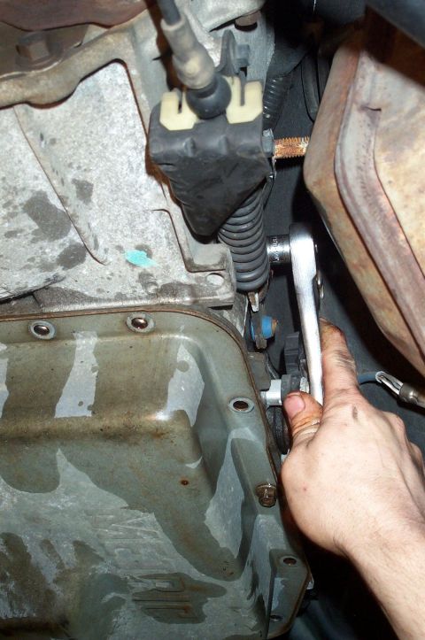 1998 Ford Crown Victoria - Underside view of a vehicle showing a hand using a ratchet wrench on linkage components near a metal oil pan and rubber boots/springs. The…
