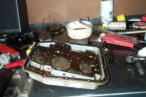 1998 Ford Crown Victoria - An automatic transmission valve body and oil pan sit on a workbench, coated in reddish-brown transmission fluid, with the gasket surface, b…