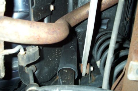 1998 Ford Crown Victoria - Underbody view showing a rusty exhaust pipe crossing above a dark rear shock absorber and a coiled black rubber hose or spring, with a moun…