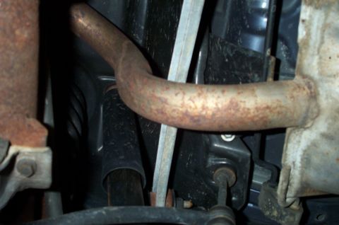 1998 Ford Crown Victoria - Close-up underside view of a vehicle chassis showing a rust-mottled curved metal exhaust or transfer pipe crossing the frame, with a black…