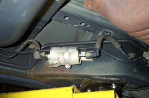1998 Ford Crown Victoria - Inline fuel filter clamped to a rusty frame rail with pressure and return lines connected, viewed from underneath the vehicle.