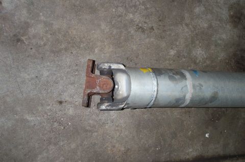 1998 Ford Crown Victoria - Close-up of a driveshaft laid on a concrete floor, showing the metal tube with faint yellow and blue paint marks and a rusty U-joint yoke f…