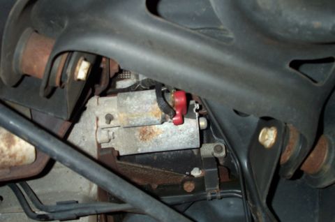 1998 Ford Crown Victoria - Underbody close-up of a rusted metal bracket and lever with a red plastic clip, surrounded by black hoses and frame.