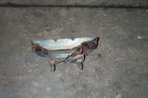 1998 Ford Crown Victoria - A rusty metal transmission crossmember or mount bracket resting on a dusty concrete floor, showing two bolt holes on the upper flanges and…