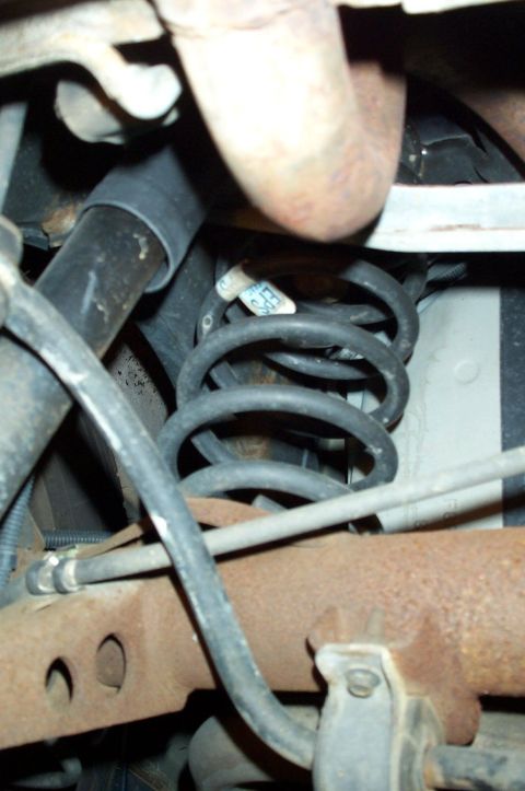 1998 Ford Crown Victoria - Underbody view of a coil suspension spring with a paper tag reading 'EPS' still attached, surrounded by a rusted exhaust pipe, brake and fu…
