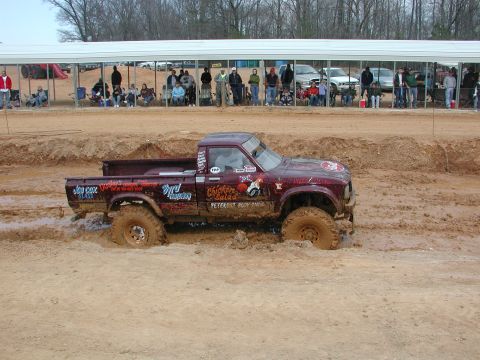 1985 Ford Bronco - A modified pickup truck navigates a muddy course during an event, with onlookers observing from a shaded grandstand.