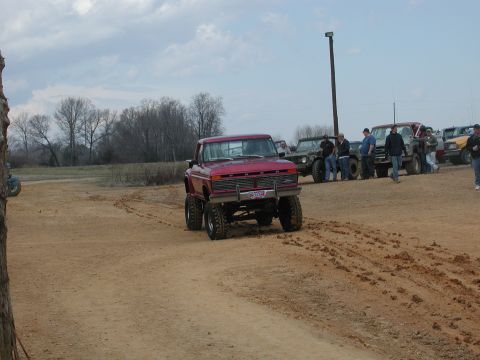 1985 Ford Bronco - Red pickup truck with lifted suspension on a dirt track, with other vehicles and people present.