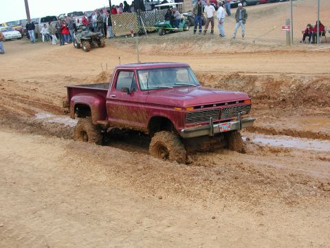 1985 Ford Bronco - A red pickup truck navigates a muddy obstacle course, with spectators in the background.