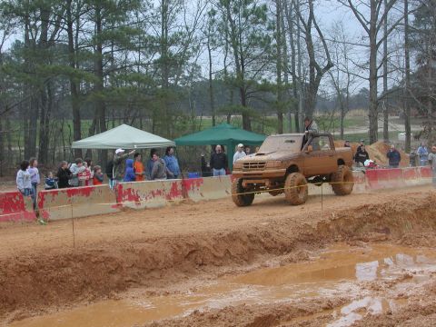 1985 Ford Bronco - A lifted pickup truck navigates a muddy track during an off-road event with onlookers present.